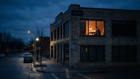 A single lit window in a dark brick office building at dusk, someone working late inside